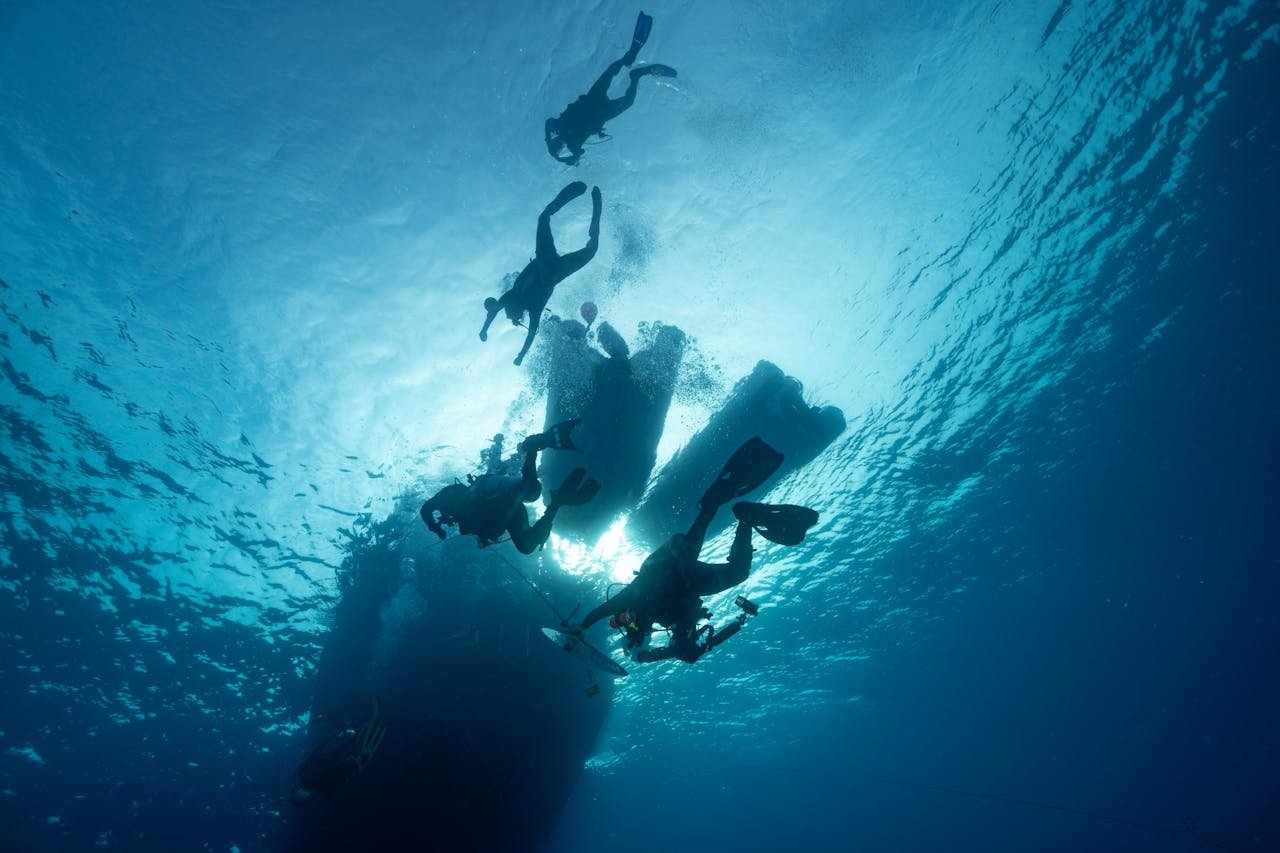 Silhouetted divers swimming beneath a yacht in clear ocean waters.